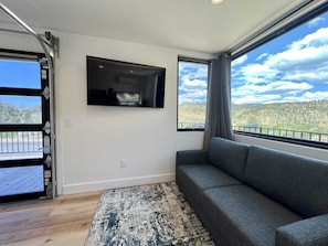 A room with a gray sofa, a mounted TV, a large window with a view of a mountainous landscape, and a wooden floor. - Modern Cabin w/Resort Pool, Hot Tubs & Pickleball (Orderville)