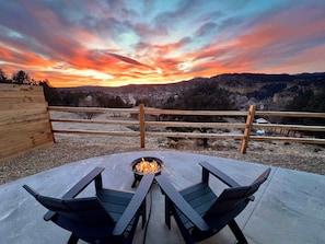 Une terrasse en bois avec un foyer et deux fauteuils Adirondack, offrant une vue sur un paysage montagneux.