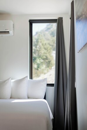 A hotel room with a bed, white pillows, a window with a view of trees, and an air conditioning unit. - Modern Cabin Near Zion & Bryce National Parks (Orderville)