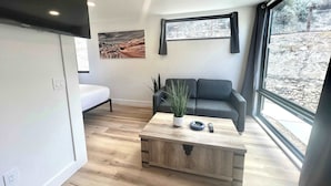 A modern living room with a sofa, a coffee table, a painting, and a large window. - Modern Cabin Near Zion & Bryce National Parks (Orderville)