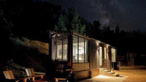Et moderne glas-hus med bålsted og udendørs siddepladser, omgivet af en stjerneryg nattehimmel.