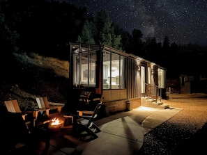 A modern glass house with a fire pit and outdoor seating, surrounded by a starry night sky.