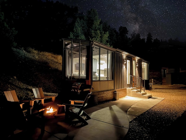 A modern glass house with a fire pit and outdoor seating, surrounded by a starry night sky.