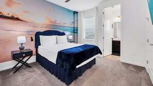 Ein Schlafzimmer mit einem Bett, einem Nachttisch, einem Deckenventilator, einem Wandbild mit einer Strandansicht und einer Tür zu einem Badezimmer.