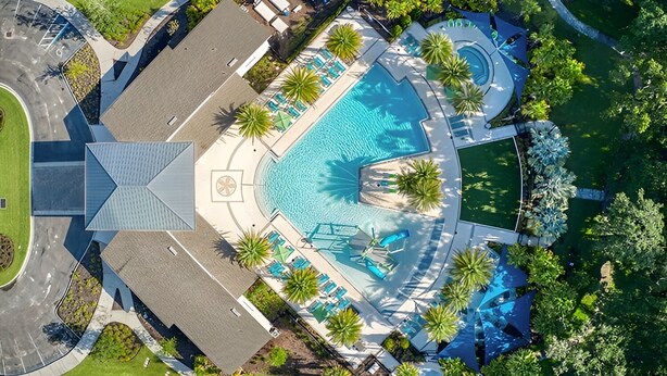 Eine Vogelperspektive zeigt ein Resort mit einem zentralen Schwimmbad, umgeben von Palmen und Liegestühlen.