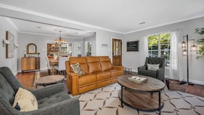A spacious living room with a brown leather sofa, a grey armchair, and a round coffee table. A dining area with a wooden table and chairs is visible in the background. - The private home features two master suites with King beds and full attached bathrooms. The loft is 3rd private getaway with a Queen bed and attached full bath. Fully stocked kitchen and a separate Oak dining area. (Dallas)