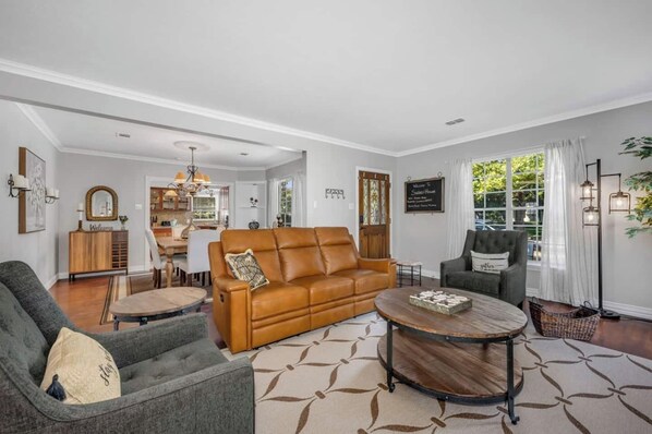 A spacious living room with a brown leather sofa, a grey armchair, and a round coffee table. A dining area with a wooden table and chairs is visible in the background.