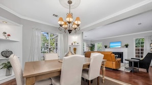 A spacious living room with a wooden dining table, white upholstered chairs, a sofa, and a TV mounted above a fireplace. - The private home features two master suites with King beds and full attached bathrooms. The loft is 3rd private getaway with a Queen bed and attached full bath. Fully stocked kitchen and a separate Oak dining area. (Dallas)