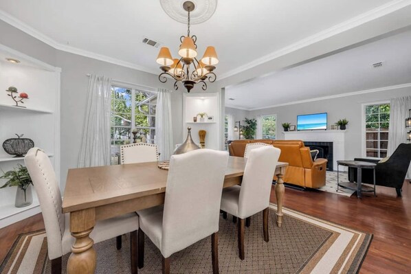 A spacious living room with a wooden dining table, white upholstered chairs, a sofa, and a TV mounted above a fireplace.