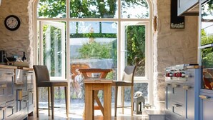 A modern kitchen with a wooden table and chairs, large windows, and a stone wall.