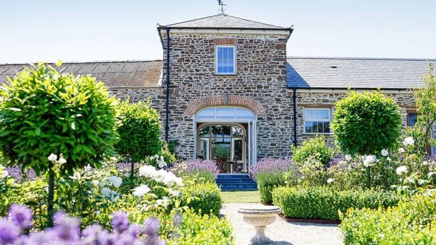 A stone building with a central entrance, surrounded by manicured gardens and a gravel path.