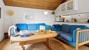 Een gezellige woonkamer met een houten plafond, een blauwe bank, een houten tafel met een spel en een keuken in de achtergrond.