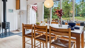 Een eethoek met een houten tafel en stoelen, een zwarte kookplaat en grote ramen met uitzicht op groen.
