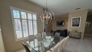 A dining area with a glass table, white chairs, a chandelier, and a kitchen in the background. - 5 Star-Rated Indian Wells 5 Bedroom, Mtn View, Cas (Indian Wells)