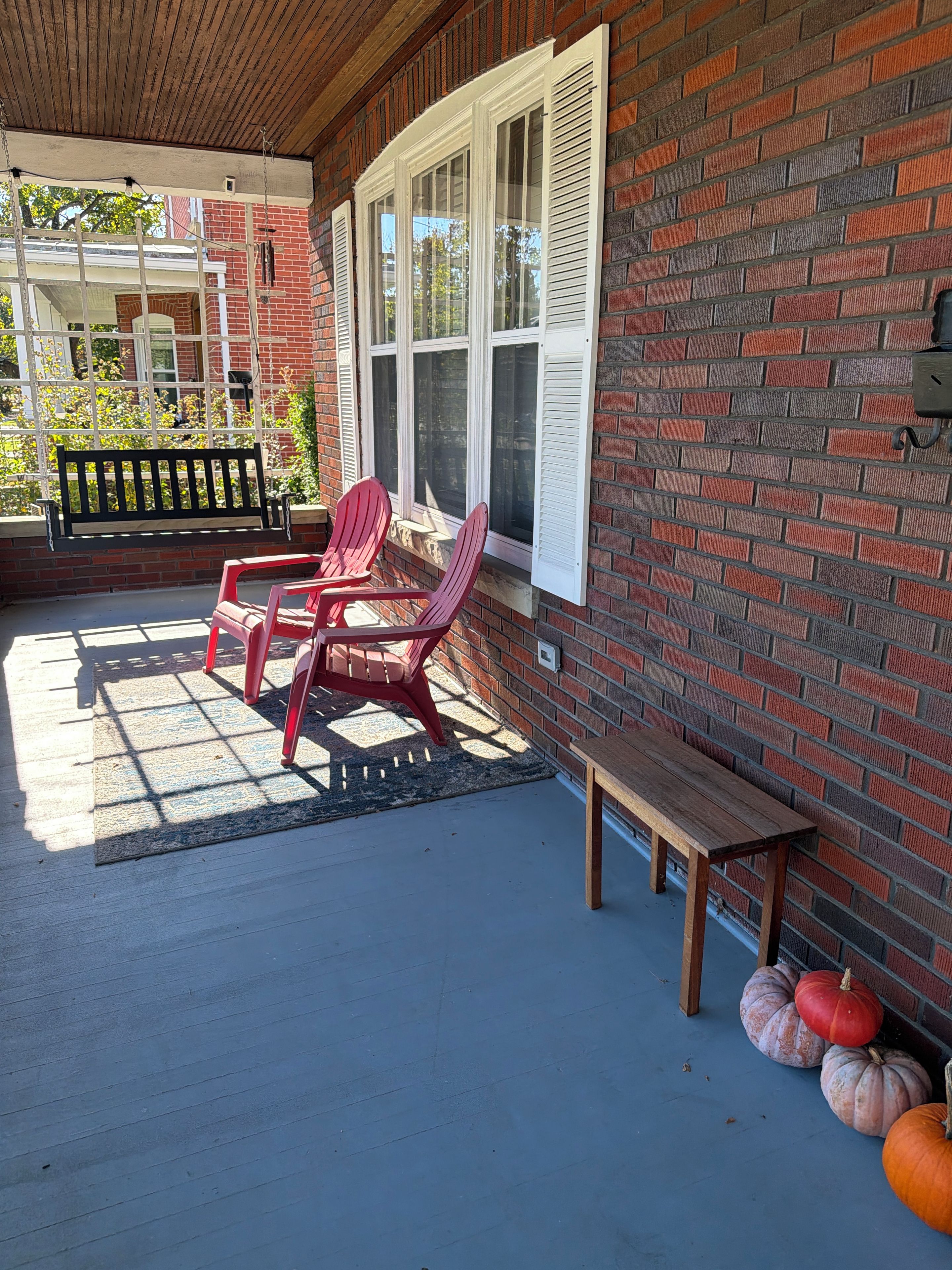 A covered porch with a brick wall, a wooden bench, two Adirondack chairs, and a small table.