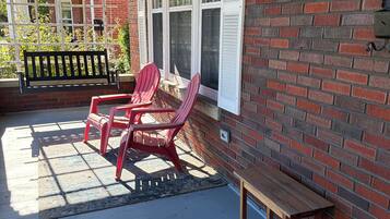 A covered porch with a brick wall, a wooden bench, two Adirondack chairs, and a small table.