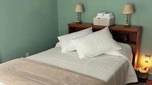 Un lit bien fait, avec du linge de lit blanc et un plaid beige, une table de chevet en bois avec une lampe et un panier, et un mur orné de deux œuvres d’art encadrées.