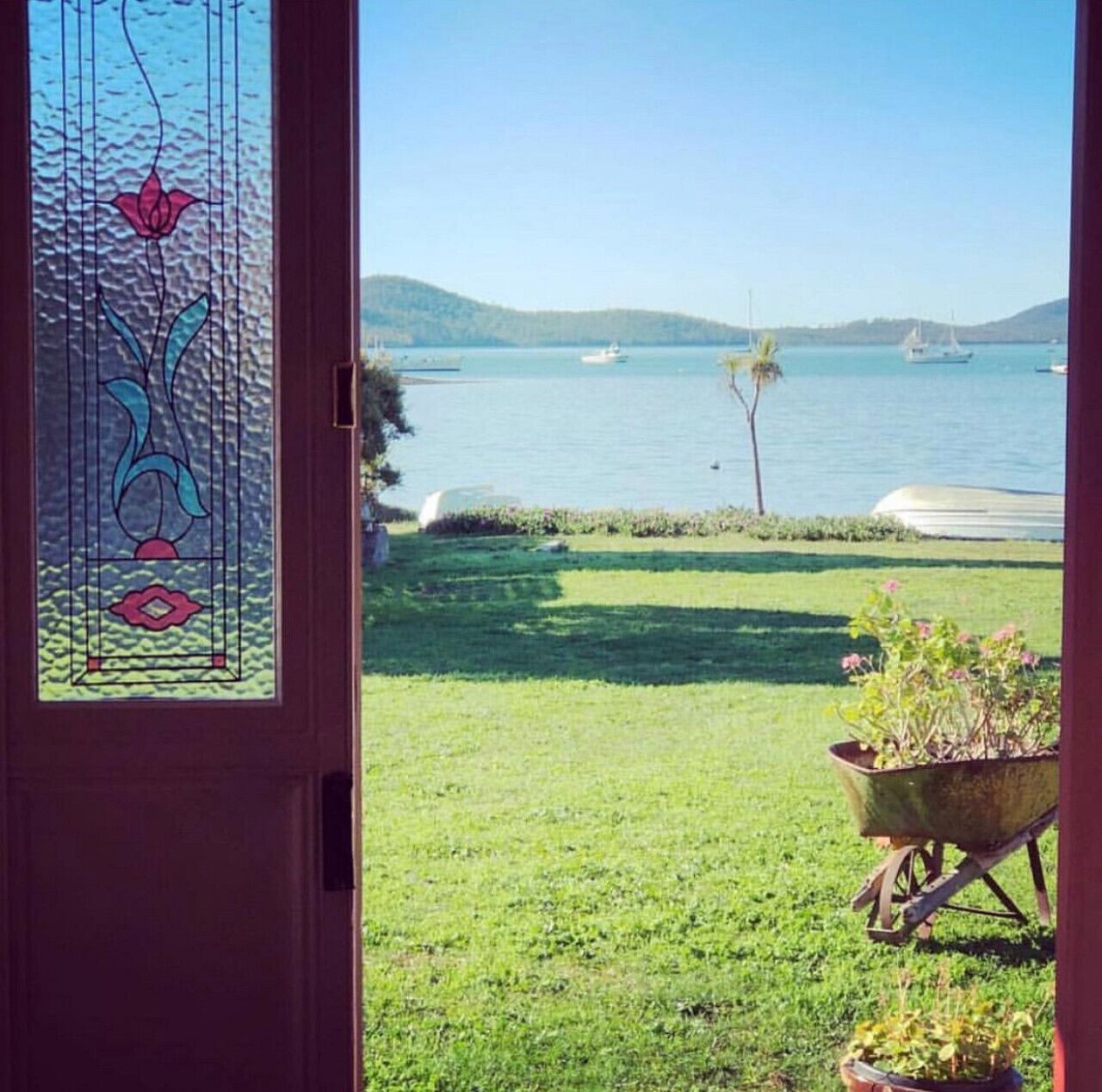 A view through a glass door with a decorative design, a grassy yard, a wheelbarrow with plants, and a body of water with boats in the distance.