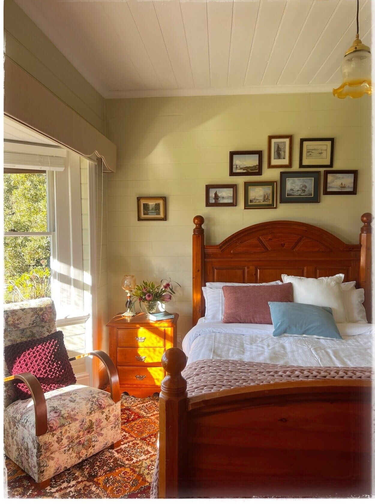 A bedroom with a wooden bed, a nightstand, a chair, and a window with a view of greenery.