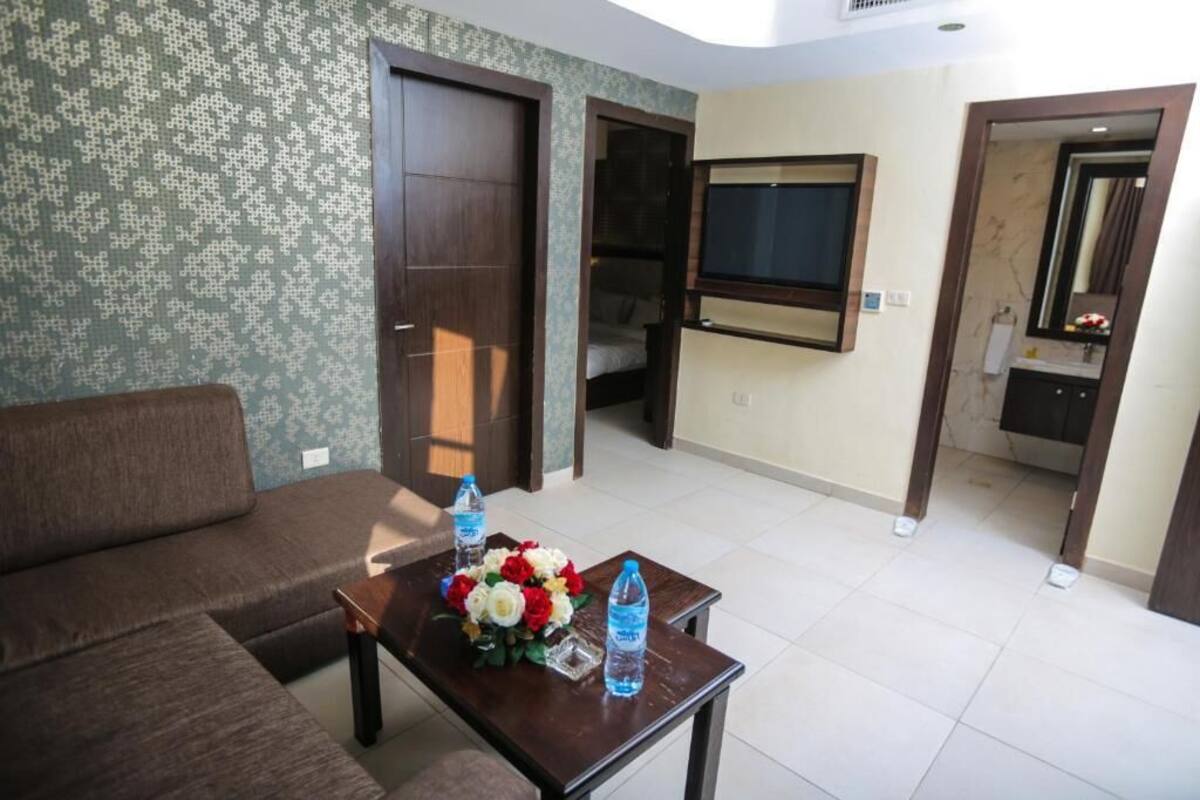 A living room with a sofa, a coffee table with flowers and water bottles, a TV mounted on the wall, and a bathroom visible through an open door.