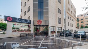 La entrada de Lancaster Hotel Apartments, con un camino despejado que lleva a la entrada, un letrero y algunos coches estacionados.