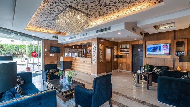 Un vestíbulo de hotel con diseño moderno, que incluye un mostrador de recepción, una zona de estar con sofás, una araña y una pantalla de televisión.