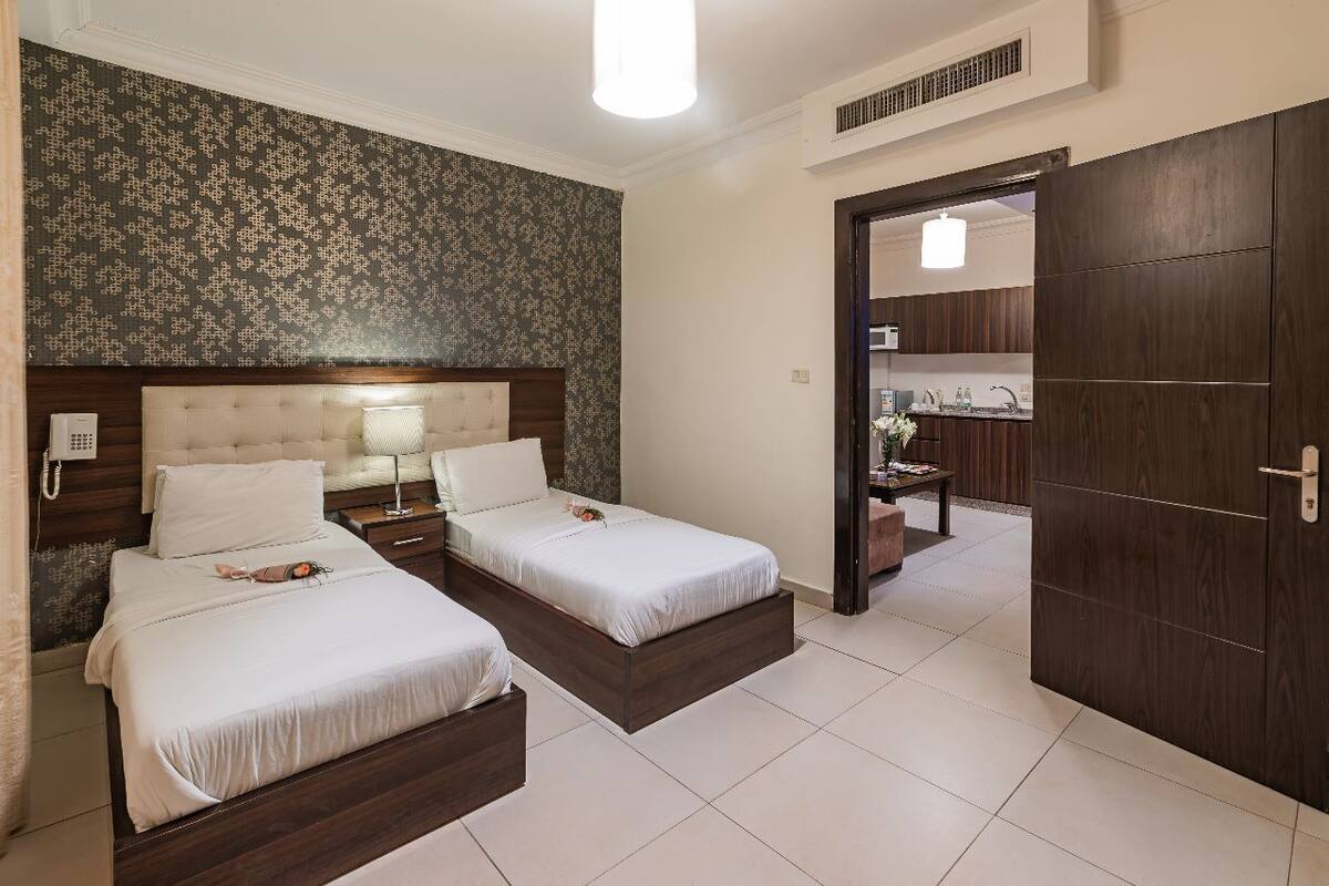 A hotel room with two beds, a wooden headboard, a bedside table with a lamp, a telephone, and a bathroom visible through an open door.