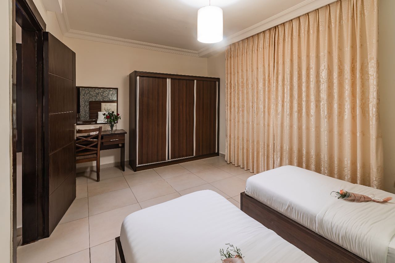 A hotel room with two beds, a desk, a wardrobe, and a large window with curtains.