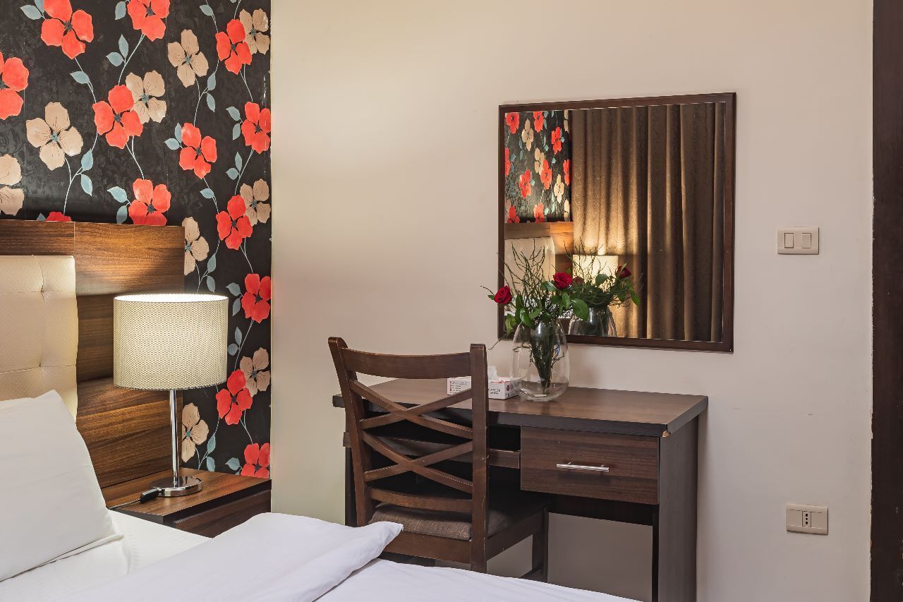 Une chambre d’hôtel comprenant un lit, un bureau, une chaise, une lampe, un miroir et un papier peint à motifs floraux.