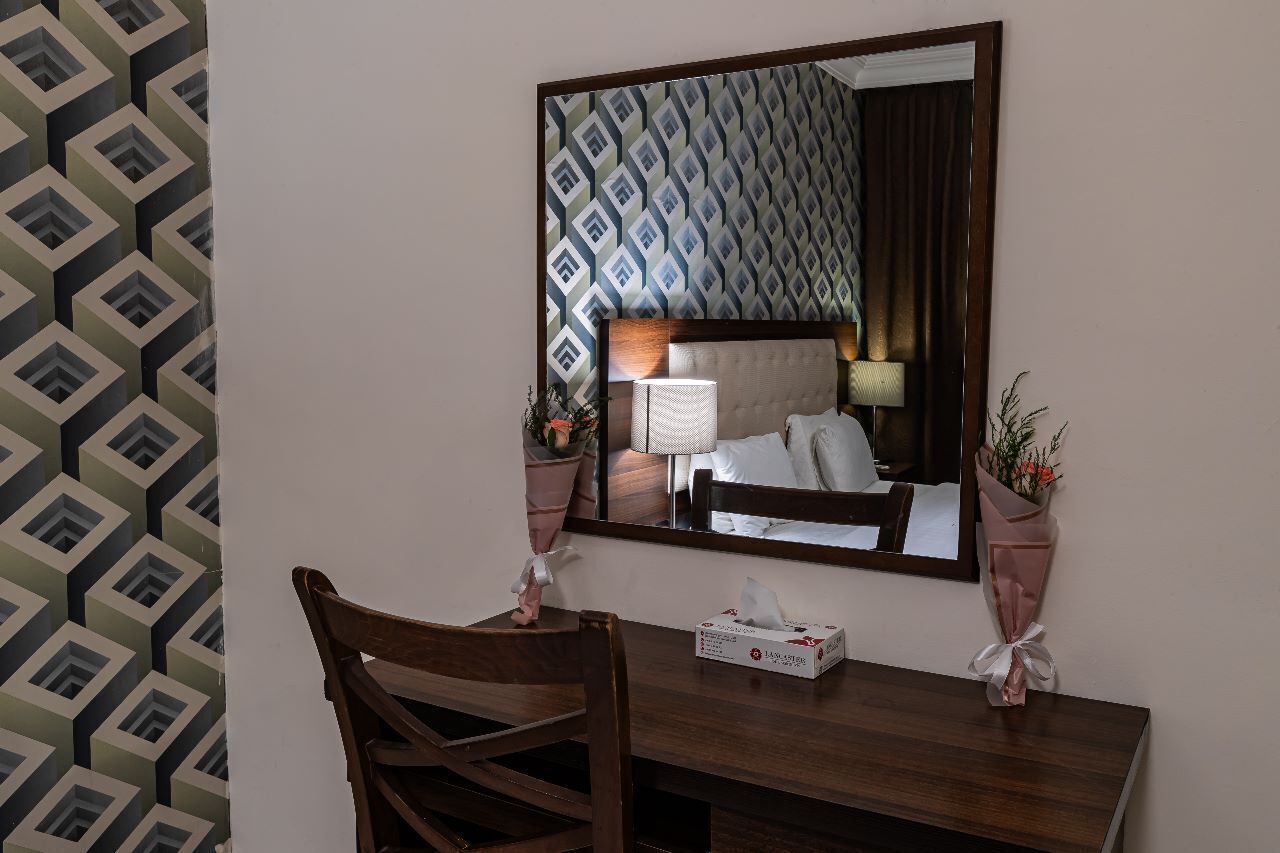 Habitación de hotel con un espejo grande, un escritorio de madera, una mesita de noche, una lámpara, un ramo de flores y un papel tapiz estampado.