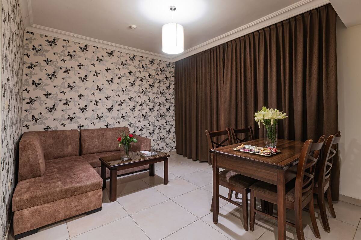 A living room with a brown sofa, a coffee table with a vase, a dining table with chairs, and a large window with curtains.