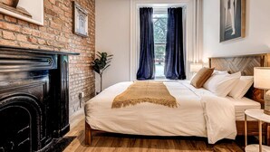 A bedroom with a brick wall, a fireplace, a bed with a beige blanket, and a window with dark curtains. - Parliament Views | Historic Stone Apartment in Old Quebec (Québec)