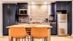 A modern kitchen with dark cabinets, a brick wall, a stainless steel refrigerator, and two orange chairs. - Parliament Views | Historic Stone Apartment in Old Quebec (Québec)
