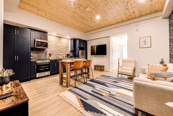 A modern kitchen with dark cabinets, a wooden ceiling, and a dining area with a striped rug.
