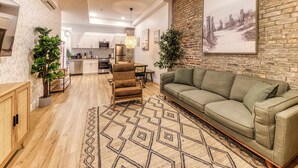 A modern living room with a green sofa, a wooden coffee table, and a patterned rug. There is a kitchen in the background with white cabinets and stainless steel appliances. - Bohemian Old Quebec Retreat | Exposed Brick & Character (Québec)