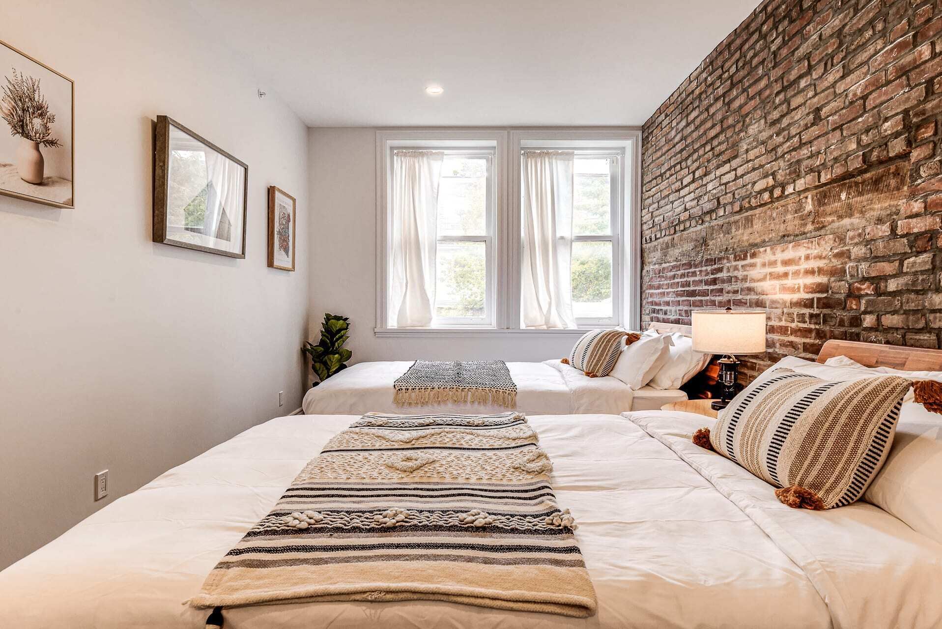 A bedroom with two beds, a brick wall, and a window with curtains.