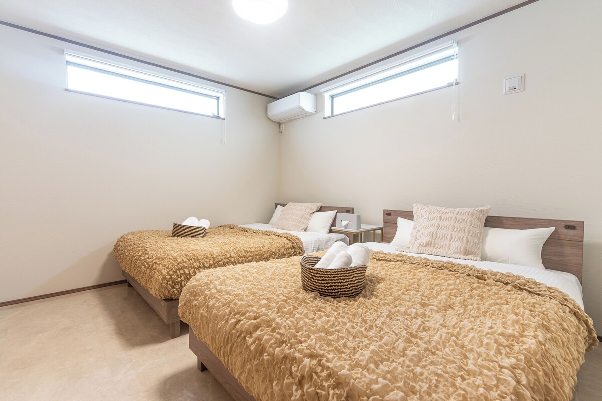 A hotel room with two beds, each with a brown textured blanket and a woven basket. There are two bedside tables with lamps, a small nightstand, and a wall-mounted air conditioning unit.