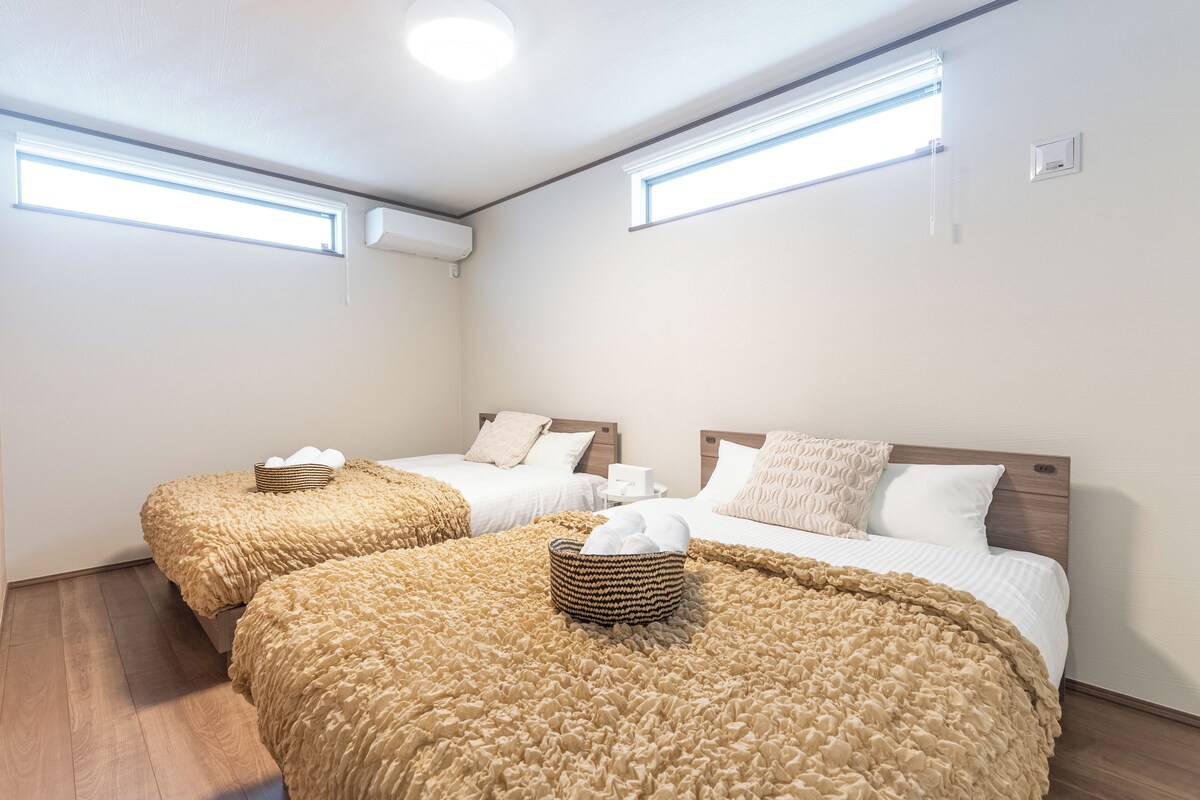 A hotel room with two beds, each with a beige textured blanket and a basket of towels. There are two windows with blinds, and the room has a wooden floor.