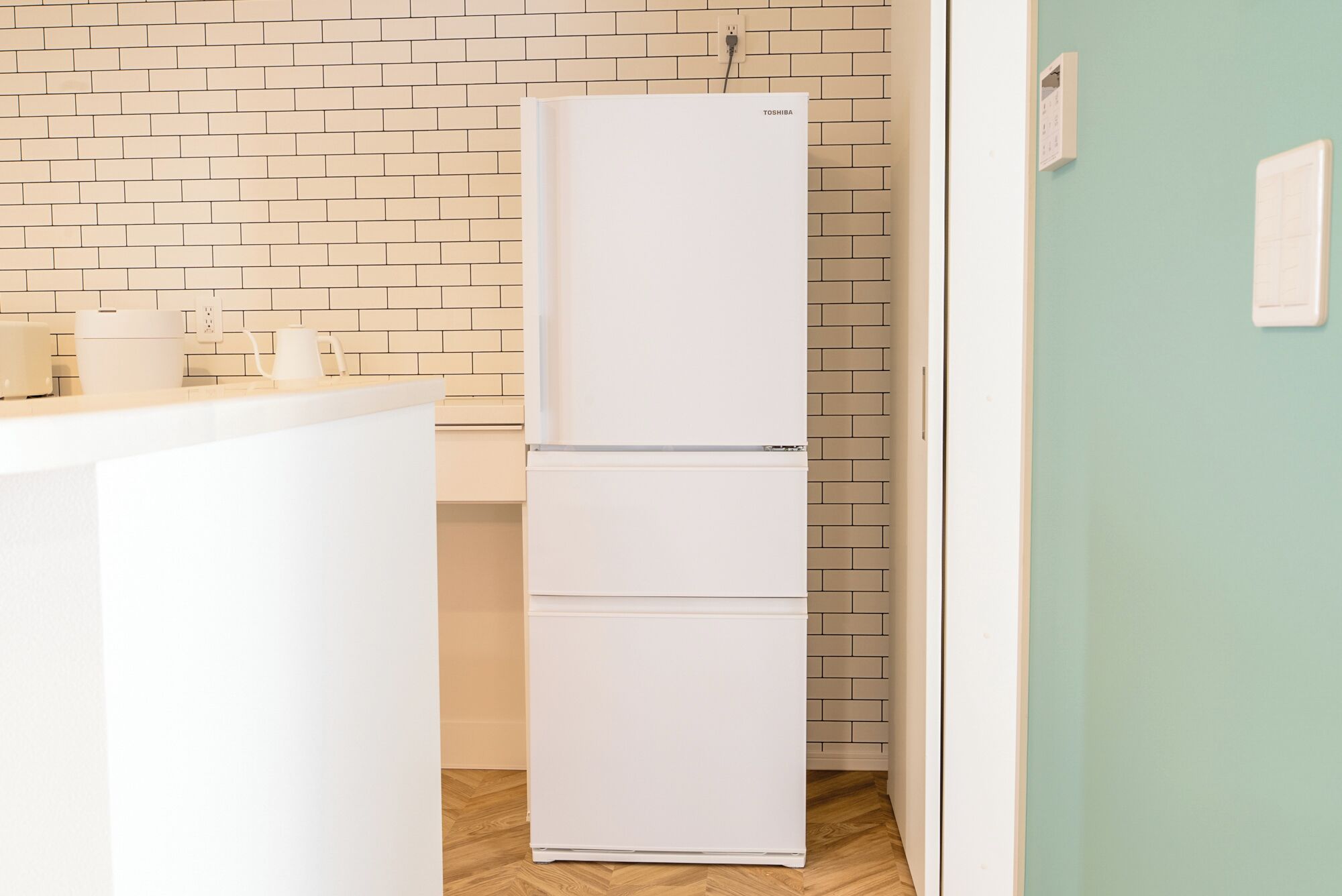 Un refrigerador blanco en una cocina con pared de ladrillo y puerta verde claro.