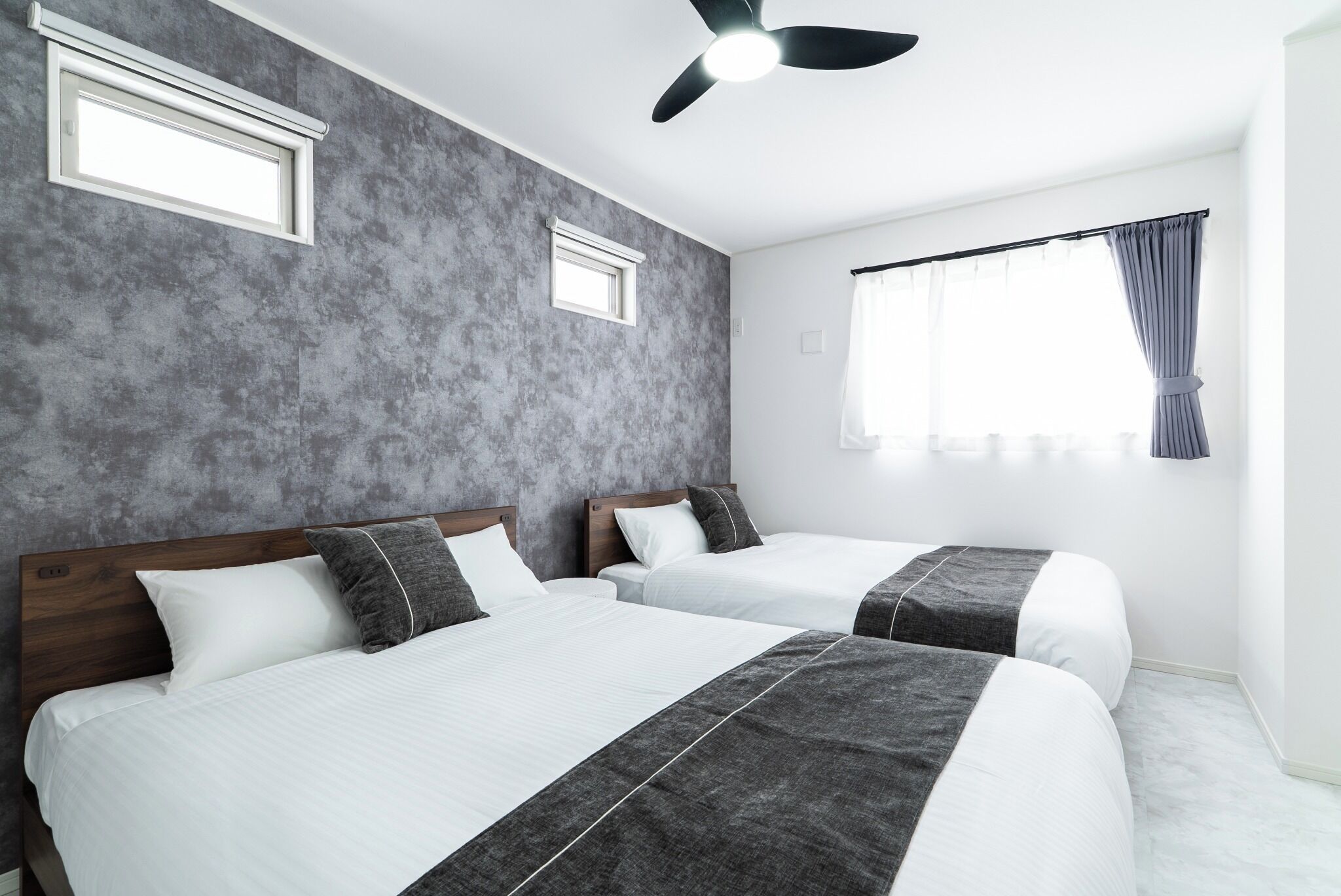 Habitación de hotel con dos camas, ventilador de techo, ventana con cortinas y pared texturizada.