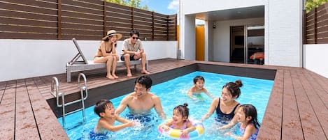 En swimmingpool med ett trädäck, en möblerad uteplats och en modern byggnad i bakgrunden.