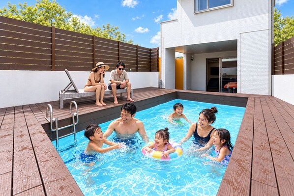 A swimming pool with a wooden deck, a covered seating area, and a modern building in the background. - Grandioso Okinawa Pool Villa Onna 8 (Onna)