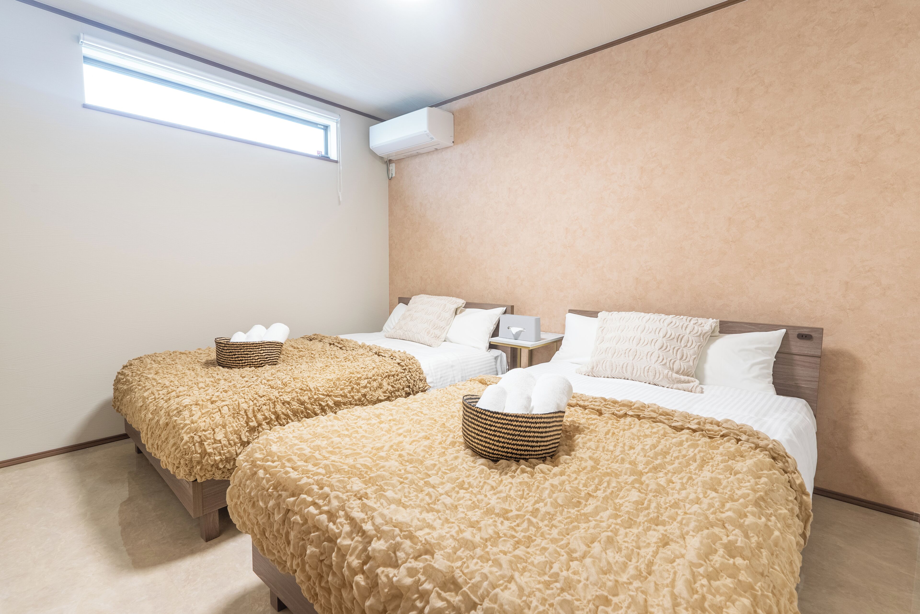 A hotel room with two beds, each with a textured beige blanket and a basket of towels.