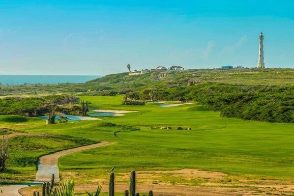 Un campo de golf con búnkeres, un estanque, un faro y un edificio a lo lejos.