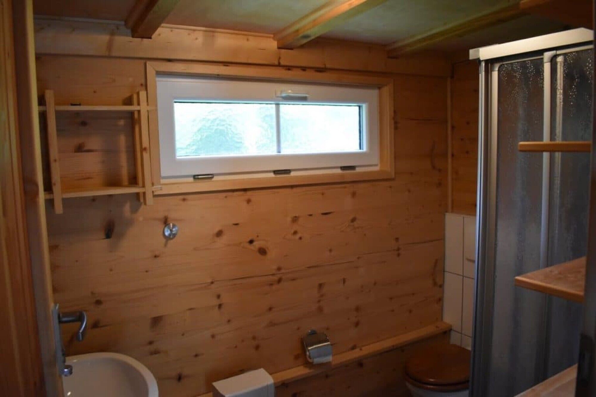 A wooden cabin bathroom with a window, a toilet, a sink, and a shower enclosure.
