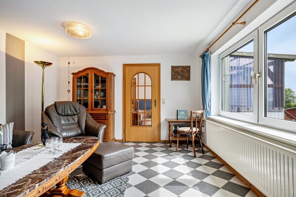 Une pièce avec un sol à carreaux, un fauteuil en cuir, une armoire en bois, une table à manger avec des chaises et une fenêtre avec des rideaux.