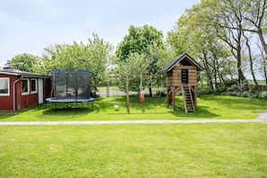 Un jardin avec un trampoline, une cabane en bois avec une échelle et un bâtiment rouge.