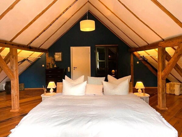 A bedroom with a sloped ceiling, wooden beams, a bed with white bedding, two bedside tables with lamps, and a door.