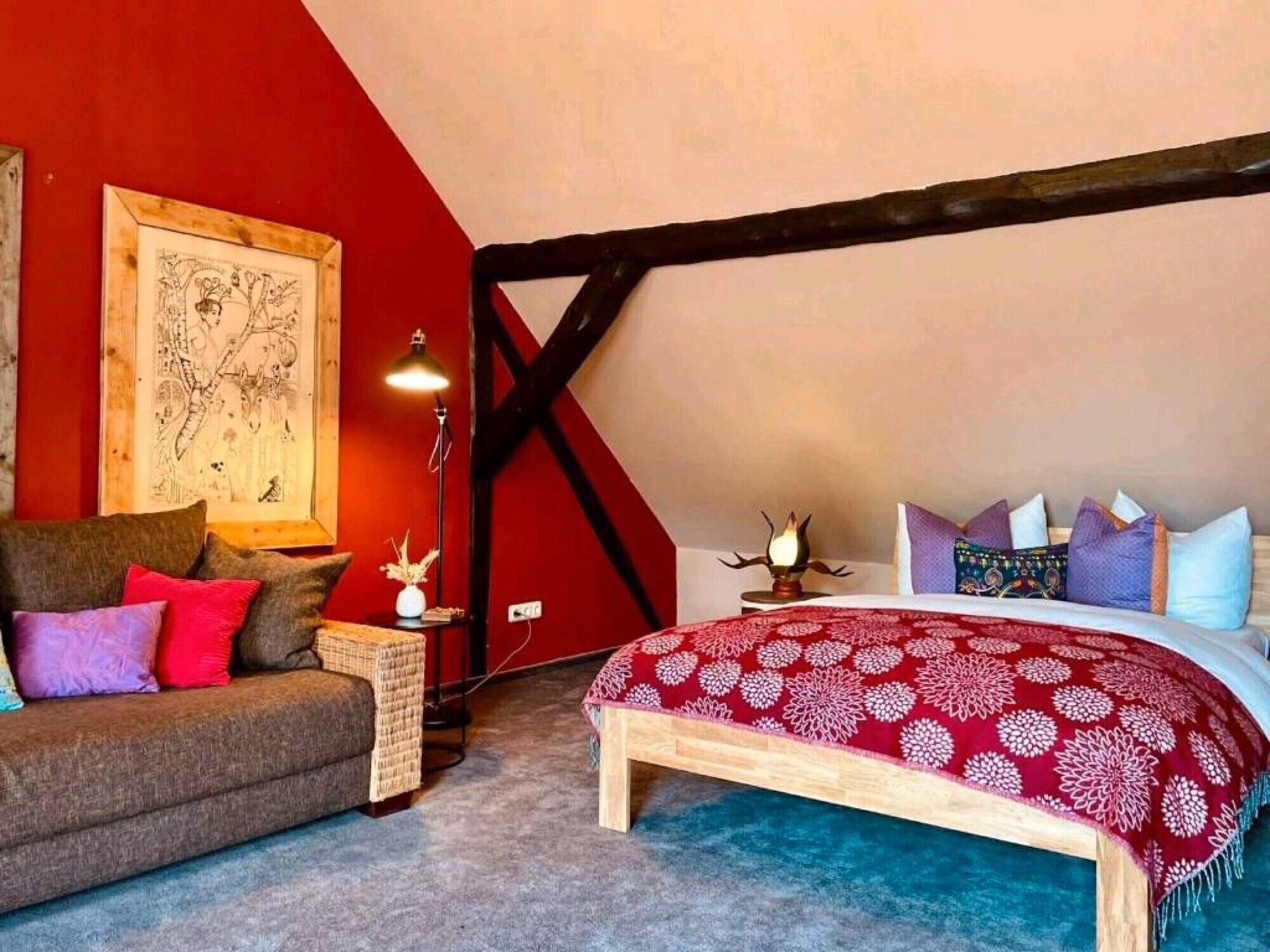Camera da letto con soffitto spiovente, un letto con copriletto floreale rosso, una testiera in legno, una parete rossa con una mappa, un divano marrone con cuscini rossi e viola, e un tavolino con una lampada e un vaso.