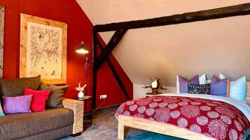 Camera da letto con soffitto spiovente, un letto con copriletto floreale rosso, una testiera in legno, una parete rossa con una mappa, un divano marrone con cuscini rossi e viola, e un tavolino con una lampada e un vaso.